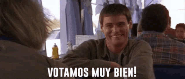 a man is smiling while sitting at a table and the words votamos muy bien are on the screen
