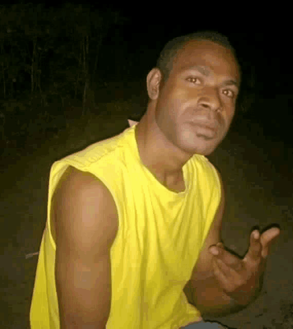 a man in a yellow tank top is making a funny face while sitting on the ground .