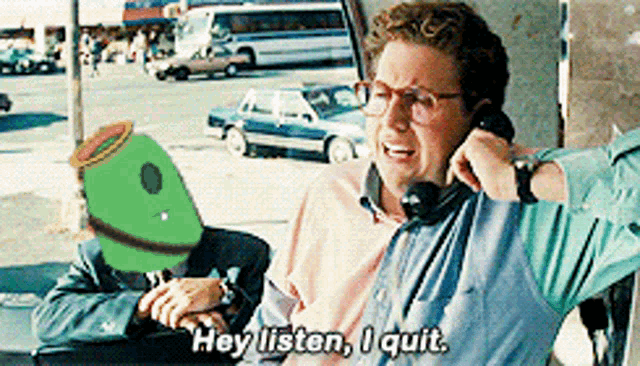 a man talking on a cell phone with a green puppet behind him saying hey listen i quit