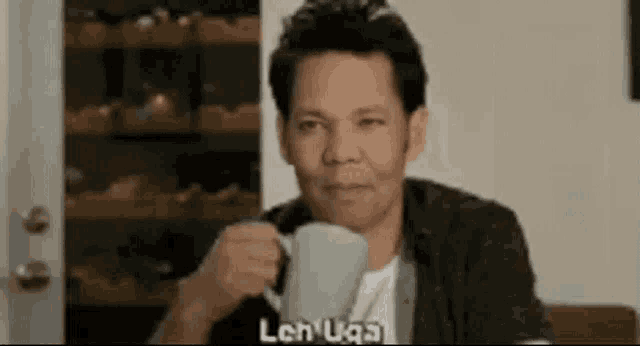 a man is sitting at a table drinking a cup of coffee and making a funny face .