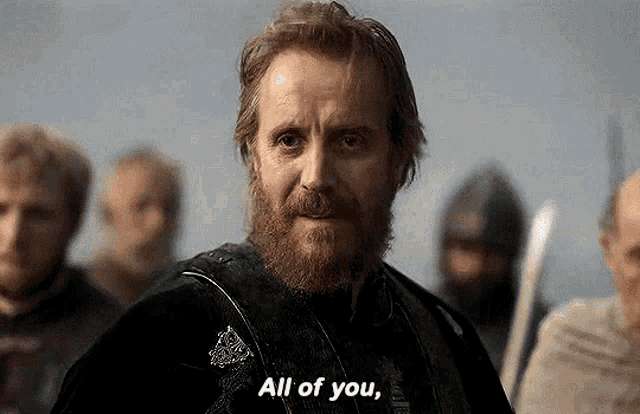 a man with a beard is standing in front of a group of people and says `` all of you '' .
