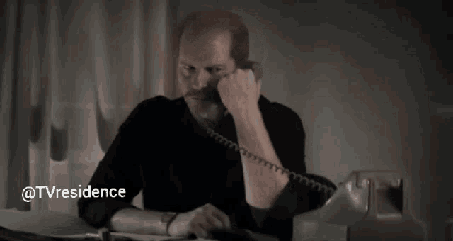 a man sitting at a desk talking on a telephone with a tv residence logo behind him
