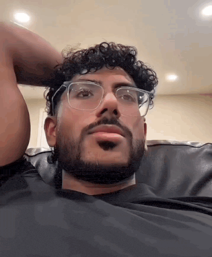 a man with curly hair and a beard wearing glasses is sitting on a couch .