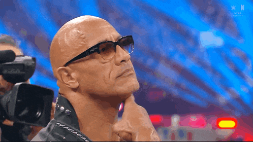 a bald man wearing glasses and a black jacket stands in front of a camera