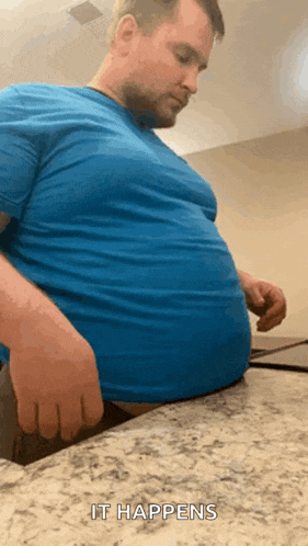 a man in a blue shirt is sitting at a counter with his hands on his belly .