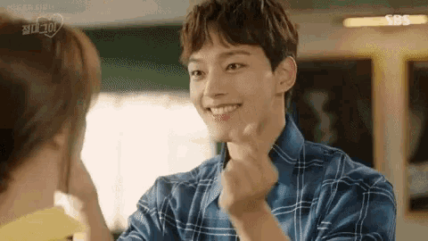 a man in a blue plaid shirt is making a heart shape with his hands while looking at a woman .