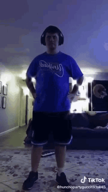 a man wearing headphones and a blue shirt with the word dallas on it stands in a living room