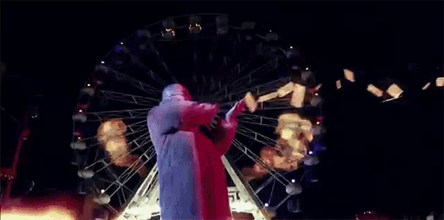 a man in a red jacket is standing in front of a ferris wheel at night .