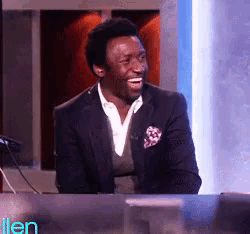a man in a suit is laughing while sitting at a desk in front of an ellen show