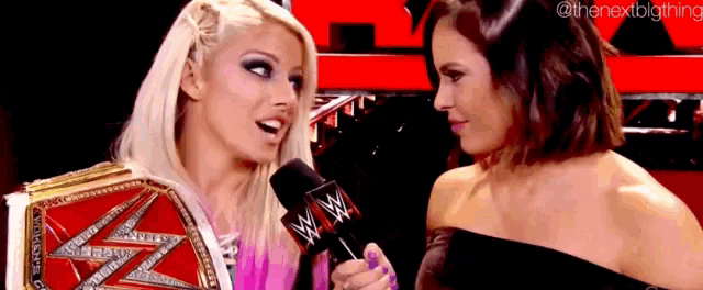 two women are talking to each other in front of a microphone . one of the women is wearing a wrestling belt .