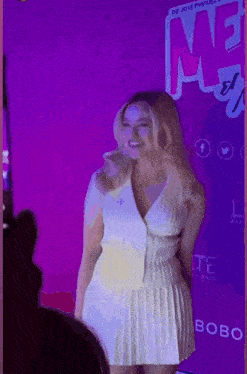 a woman in a white dress is standing in front of a purple wall with the word me on it .