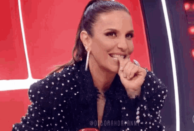 a woman in a black jacket with pearls on it is smiling and holding her finger to her mouth .
