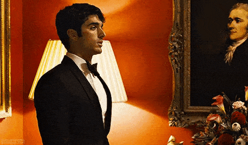 a man in a tuxedo and bow tie is standing in front of a painting of a man .