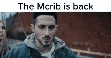 a man with a beard is standing in front of a red building with the words `` the mcrib is back '' .
