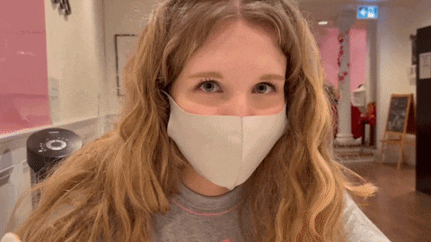 a woman wearing a white face mask looks at the camera in a hallway