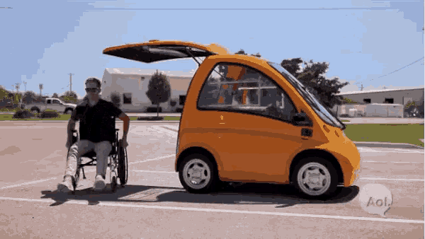 a man in a wheelchair is standing next to a small orange car