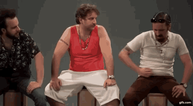 a man in a red tank top is sitting on a box with two other men .