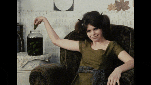 a woman in a green dress is sitting in a chair with a jar of pickles behind her