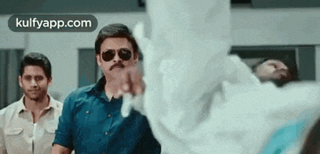 a man wearing sunglasses and a mustache is pointing at something while standing next to another man .