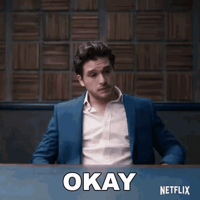 a man in a suit and pink shirt is sitting at a desk with the word okay written on it .