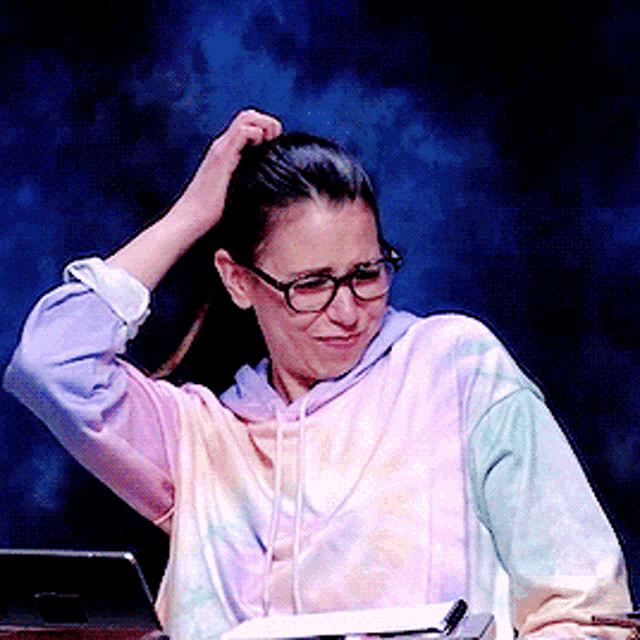 a woman wearing glasses and a tie dye sweatshirt scratches her hair