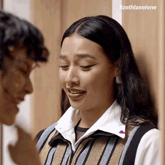 a woman in a striped vest talks to a man in a white shirt with the name izethlaselene on the bottom