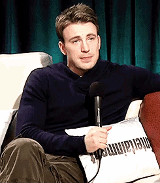 a man is sitting on a couch holding a microphone and a pillow with entertainment written on it