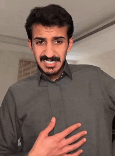 a man with a mustache is wearing a gray shirt and making a funny face with his hand on his chest .