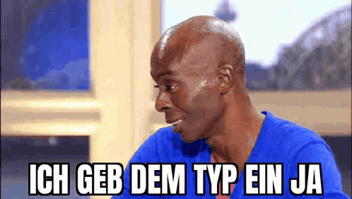 a bald man in a blue shirt is sitting in front of a window and making a funny face .