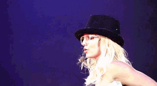 a blonde woman wearing a black hat and glasses waves her hand