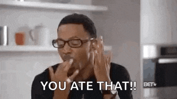 a man wearing glasses is eating something in a kitchen and a woman is holding his hand over his mouth .