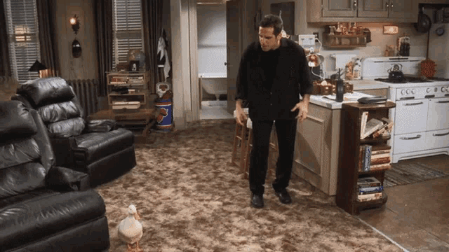 a man in a black suit stands in a living room with a duck in the foreground