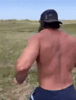 a shirtless man wearing a baseball cap is running in a field .