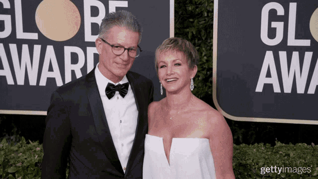 a man in a tuxedo and a woman in a white dress pose on a red carpet in front of a globe award sign