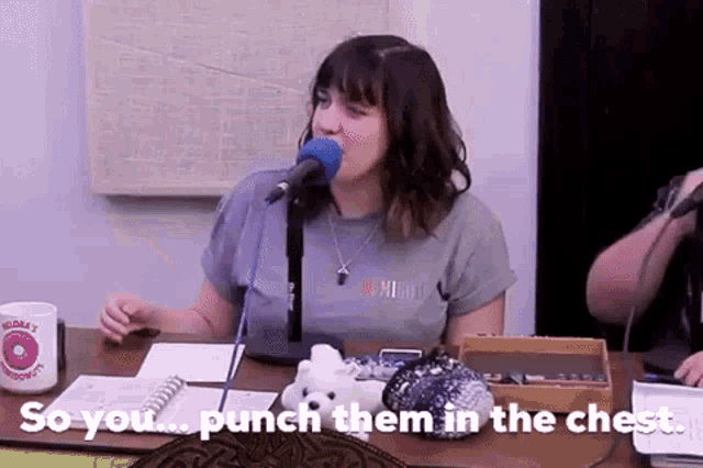 a woman is sitting at a table with a microphone in her mouth and the words `` so you punch them in the chest '' .