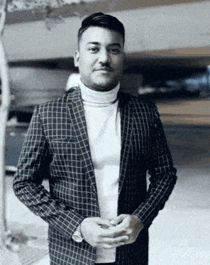 a man wearing a plaid jacket and a white turtleneck is standing with his hands folded