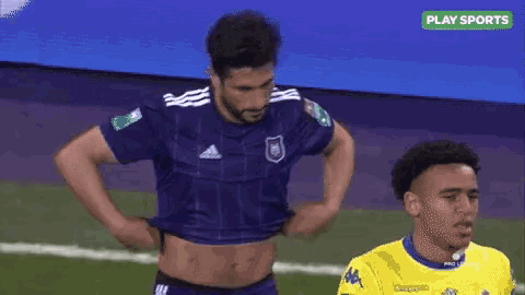 a man is taking off his shirt on a soccer field while another man looks on .