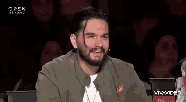 a man with a beard is sitting in front of a microphone on a stage and smiling .