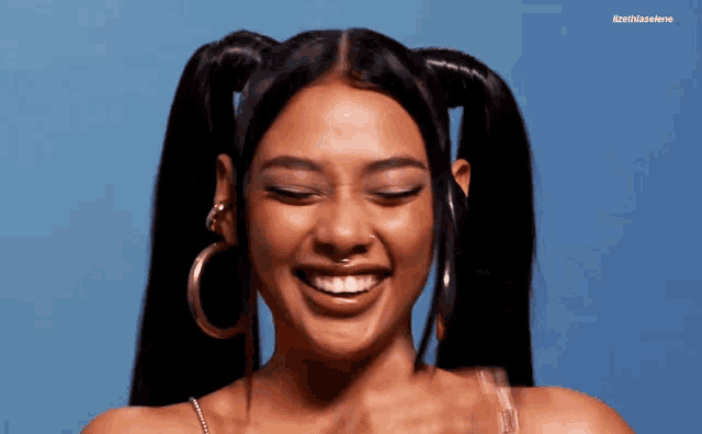 a woman with pigtails and hoop earrings smiles for the camera with a blue background behind her that says iznrthisalene