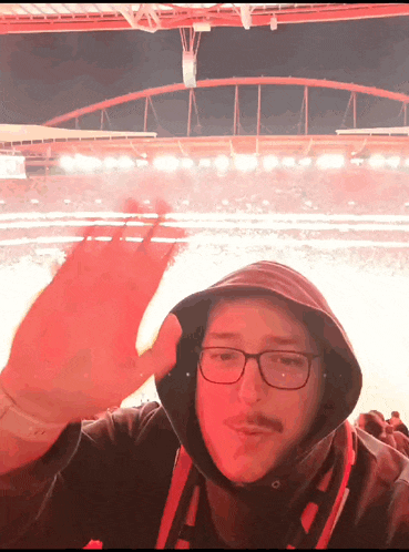 a man wearing glasses and a hooded jacket waves his hand in front of a stadium
