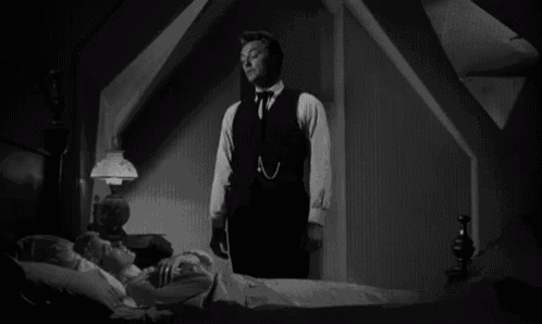 a black and white photo of a man standing next to a woman laying in a bed