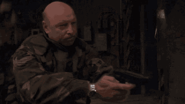 a bald man in a camouflage uniform holds a gun