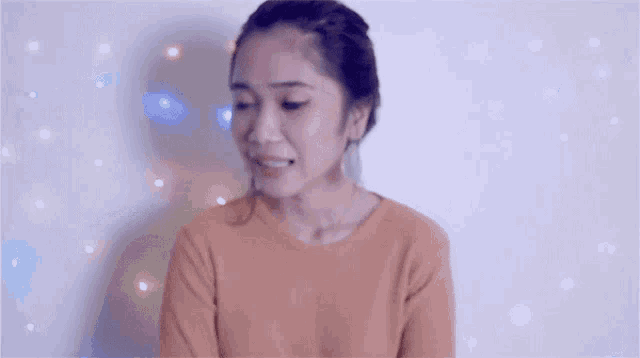 a woman in an orange sweater is standing in front of a white wall with christmas lights behind her .