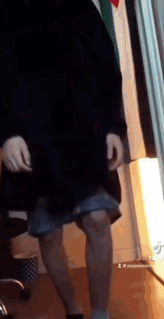 a man in a black jacket and shorts is standing in a room with his legs crossed .