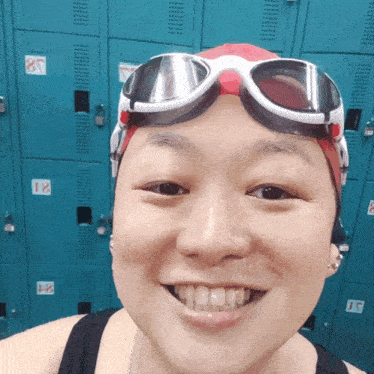 a woman wearing swimming goggles and a red swim cap smiles in front of lockers that have the numbers 18 and 19 on them