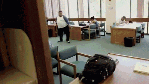 a group of people are sitting at tables in a library with a backpack on a desk .