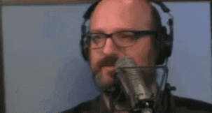 a man with glasses and a beard is talking into a microphone .