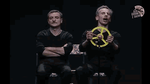 two men are sitting on a stage and one of them is holding a yellow wheel