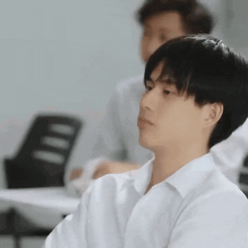 a young man in a white shirt is sitting in a classroom with other students .