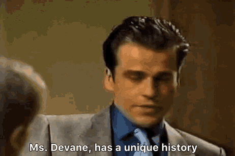 a man in a suit and tie is talking to another man and says ms. devane has a unique history .